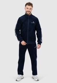 Navyblauw velours trainingspak met witte strepen, ritssluiting jack en elastische manchetten. Heeft een logo op de borst en het broekbeen.