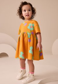 Vestido de algodón naranja con un patrón texturizado, que presenta flores azules y moradas. Se lleva con zapatillas blancas de caña alta y calcetines rosa hasta el tobillo.