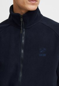 Marineblauwe fleecejack met een hoge kraag, voorzien van een ritssluiting aan de voorkant en een klein geborduurd logo op de borst. Zachte textuur en casual pasvorm.