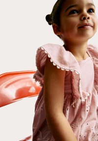 Robe en lin rose avec des manches scallopées et une broderie florale, associée à une chaise transparente rouge vif en arrière-plan.