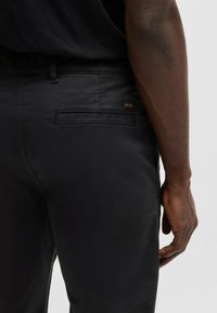 Pantaloni negri cu o textură netedă, având un buzunar posterior cu un capac rectangular și un mic logo metalic de marcă.