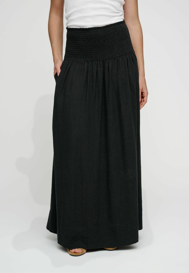 Femme portant une longue jupe noire avec une large ceinture smockée, associée à un haut blanc et des sandales, les mains reposant près des poches.