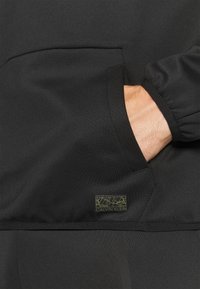 Pullover nero con tasca laterale, polsini elasticizzati e finitura in tessuto goffrato. Include un piccolo etichetta verde con logo Calvin Klein.