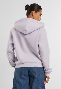 Vista posteriore di una felpa lavanda con un ampio cappuccio, polsini e orlo a coste. Il tessuto appare morbido, con una vestibilità comoda e dettagli minimali.