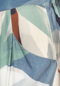 Gros plan d’un tissu avec un motif géométrique abstrait en bleu, vert, crème et marron, avec une couture et un détail de poche cousus.