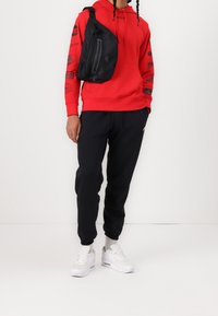 Felpa rossa con stampa grafica, borsa a tracolla nera, pantaloni sportivi scuri e sneakers bianche con dettagli grigi. Tessuto morbido e design informale.