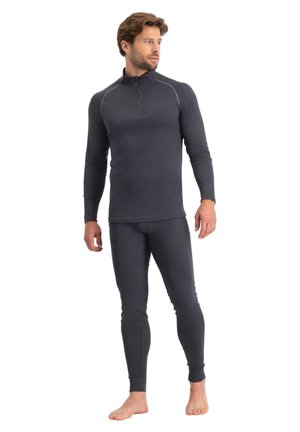 Man staat op blote voeten en draagt een nauwsluitende donkergrijze top met lange mouwen en een kwart ritssluiting, gecombineerd met bijpassende thermische leggings.