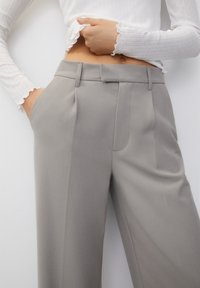 Pantalons gris sur mesure en tissu lisse, avec des plis à l'avant, des poches latérales et une taille plate. Agrémentés de détails de couture subtils.