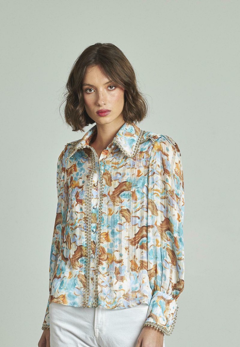 Blusa a maniche lunghe con un motivo astratto blu chiaro, bianco e marrone. Presenta un grande colletto e dettagli decorativi con bottoni.