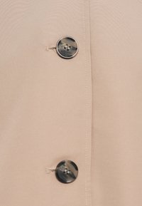 Zwei schwarz-braun marmorierte Knöpfe, die an beigem Stoff mit sichtbarem Nähfaden und Knopflöchern befestigt sind.