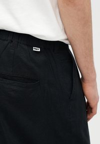 Κοντινό πλάνο ενός ατόμου που φοράει μαύρο παντελόνι με ετικέτα OBEY στη μέση και λευκό πουκάμισο μέσα από το παντελόνι.