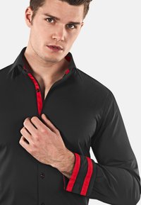 Chemise noire avec un accent rouge sur le col et les poignets. Le tissu est doux, avec une coupe ajustée et une fermeture boutonnée à l'avant.