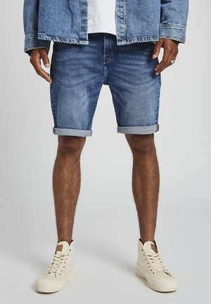 Homme portant un short en jean bleu retroussé, une veste en jean, un t-shirt blanc et des baskets montantes blanches, debout devant un fond uni.