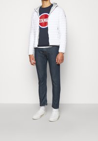 Vit puffjacka med huva, dragkedja fram, bärs över en marinblå t-shirt med en röd cirkulär logotyp, ihop med mörka jeans och vita sneakers.