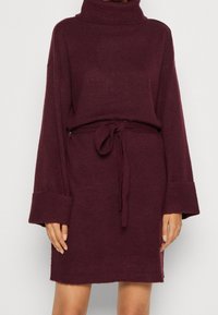 Burgunderröd stickad klänning med hög hals, vida ärmar och knut i midjan. Mjuk textur och avslappnad passform med ribbade muddar och fåll.