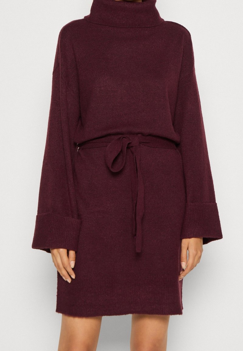 Burgunderröd stickad klänning med hög hals, vida ärmar och knut i midjan. Mjuk textur och avslappnad passform med ribbade muddar och fåll.