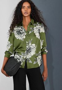 Zelená kvetinová blúzka zo saténovej látky s čierno-bielymi kvetinovými vzormi, s gombíkmi vpredu a vyhrnutými rukávmi, doplnená o čiernu kabelku s textúrou.