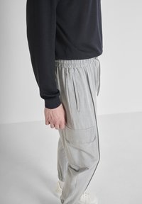 Man in lichte grijze joggingbroek met zijzakken, zwart longsleeve shirt en witte sneakers, staand voor een effen witte achtergrond.