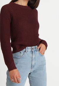 Bordowy sweter dziany o prążkowanej fakturze i dekolcie w szpic, zestawiony z jasnoniebieskimi, wysokim stanem dżinsami, posiadającymi boczne kieszenie.