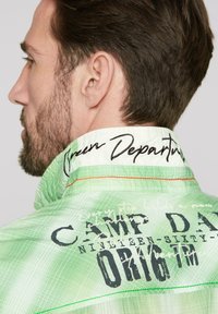 Chaqueta verde con un patrón texturizado, que presenta detalles bordados en el collar y texto impreso en la parte posterior, destacando "DÍA DEL CAMPAMENTO" y "SALIDA VERDE".