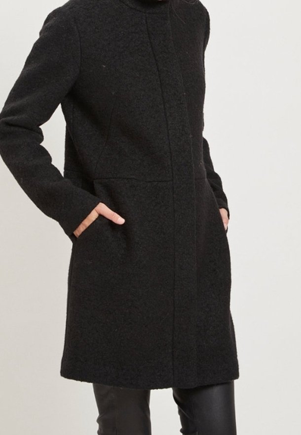 Femme portant un manteau en laine noir mi-long avec des poches latérales, debout les mains dans les poches, devant un fond clair uni.