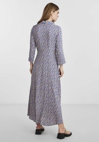 Naine seisab sinises ja beežis lillelises pikade varrukatega kleidis, beežides kontsakingades, seljaga kamera poole, lihtsa tausta ees.