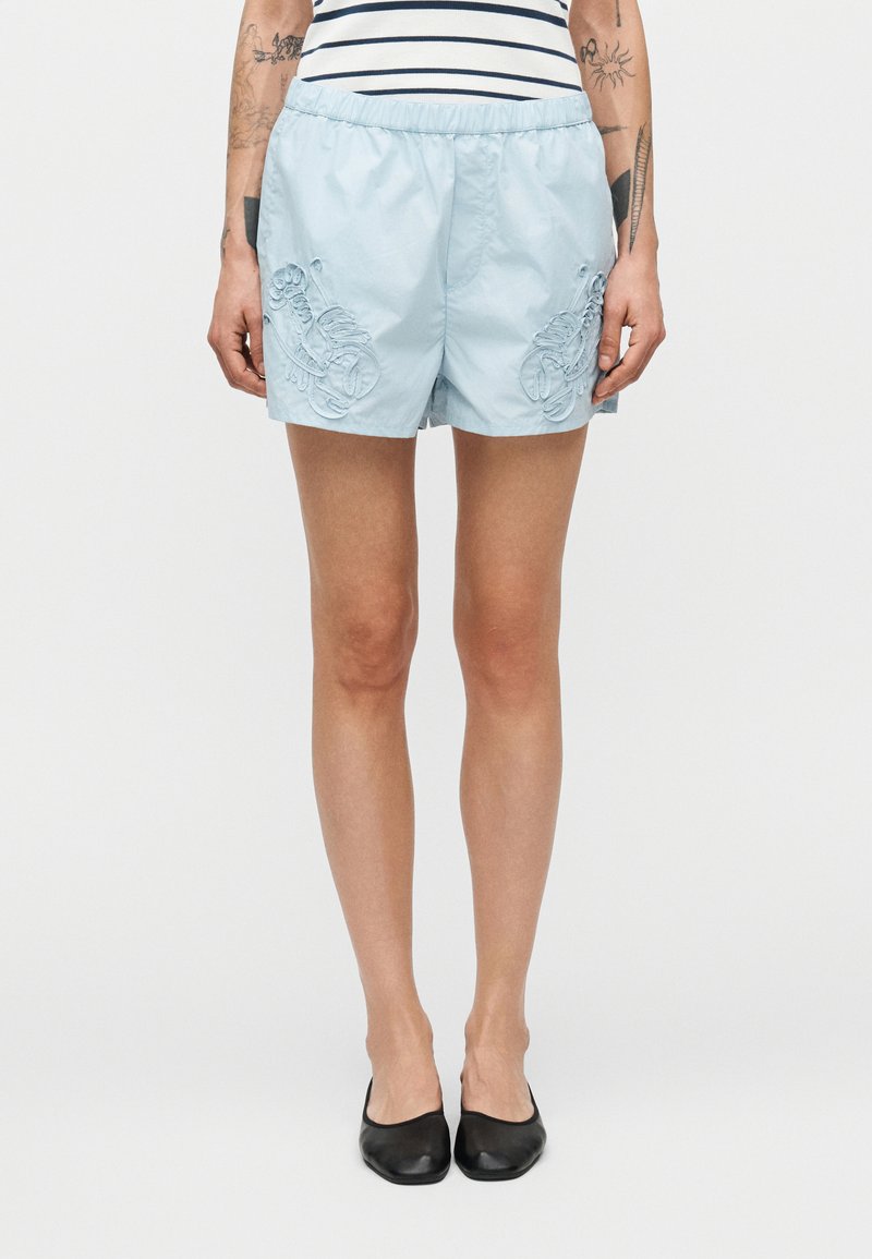 Ανοιχτό μπλε σορτς με ελαστική μέση, διακοσμημένο με κεντητά λουλουδένια σχέδια στα πλάγια. Φορεμένο με μαύρα άνετα παπούτσια.