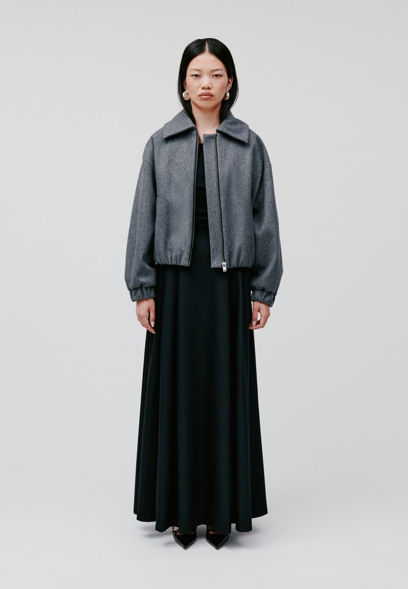 Graue Bomberjacke mit hohem Kragen, Reißverschluss und elastischen Bündchen, kombiniert mit einem langen schwarzen Kleid und spitzen schwarzen High Heels.