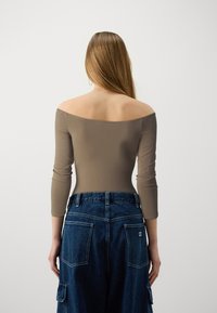 Ruda off-the-shoulder bodysuit su ilgomis rankovėmis, derinamas su tamsiai mėlynomis plačiomis džinsinėmis kelnėmis, kurių kišenės yra didelės, o ant jų yra logotipo etiketė.