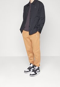 Chaqueta negra, camisa gris, pantalones de chándal beige, zapatillas de caña alta blancas y negras. La chaqueta tiene un logo en el pecho; los pantalones tienen un logo en la pierna.