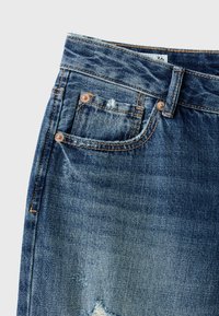 Gros plan sur un jean en denim bleu montrant une poche avant, une boucle de ceinture, des rivets en cuivre et des détails de tissu usé sur un fond clair.