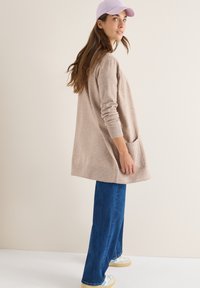 Beige oversize svetr s dlouhými rukávy a kapsami, kombinovaný s modrými širokými džíny a světle fialovou čepicí, na neutrálním pozadí.