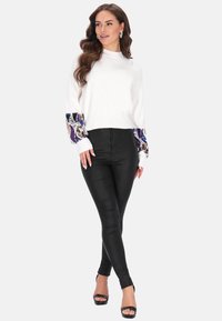 Weißer Pullover mit strukturierten, bunten gemusterten Ärmeln; kombiniert mit schwarzen, enganliegenden Hosen und schwarzen, hochhackigen Sandalen.