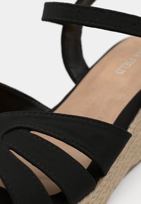 Sandale compensée noire à brides avec semelle intérieure beige et talon plateforme style espadrille tressée, gros plan sur les brides et la boucle de cheville.