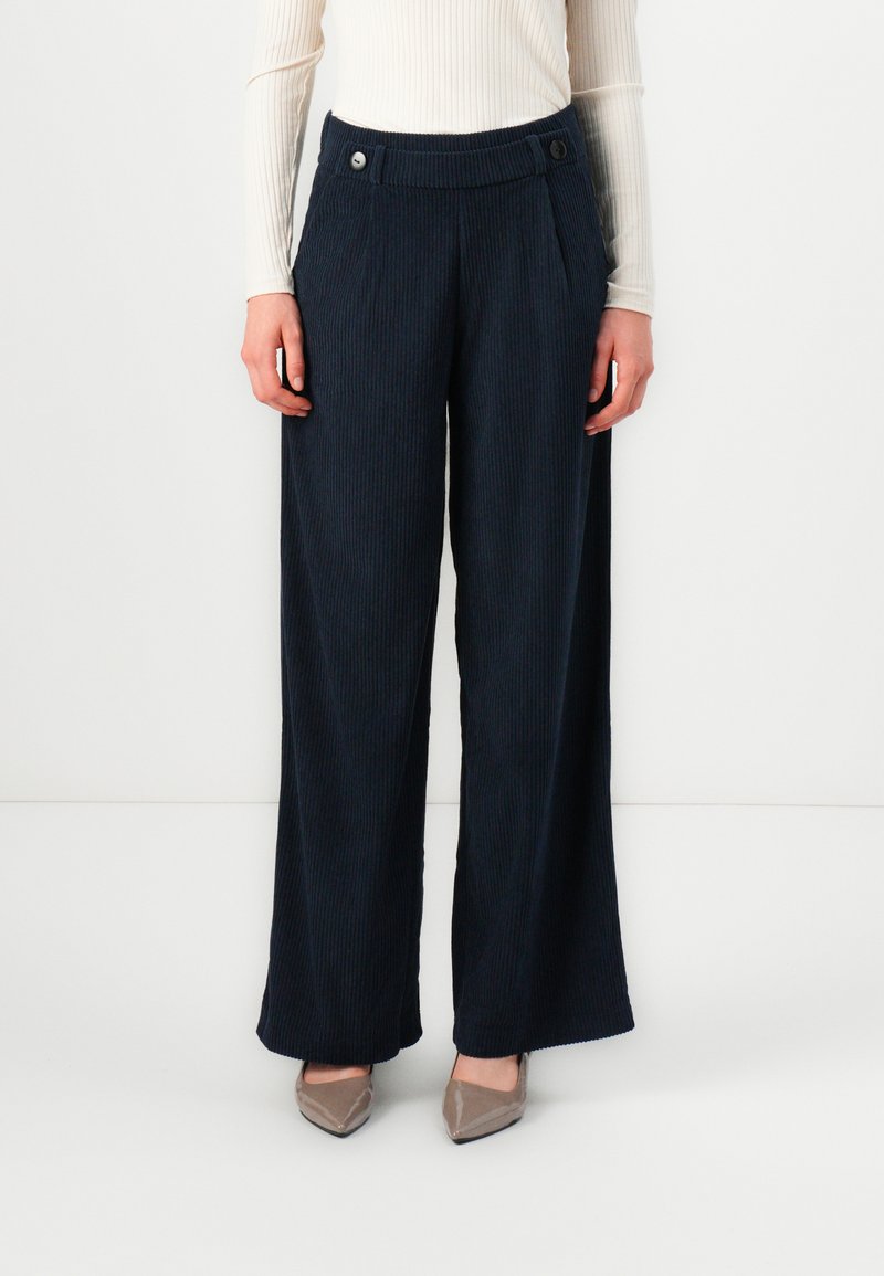 Navy corduroy wide-leg trousers with front pleats, two buttons at the waistband, and a textured surface, paired with beige pointed flats.