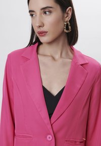 Blazer fucsia con rever a punta, chiusura con un bottone e tasche frontali. Abbinato a una maglietta nera e orecchini a cerchio dorati.