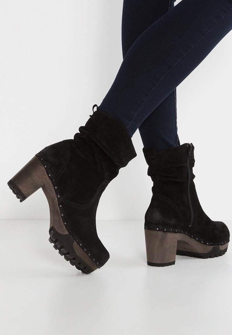 Bottines en suède noir avec un design décontracté, un talon en bois épais et des clous en métal décoratifs le long de la semelle. Fermeture éclair sur le côté.