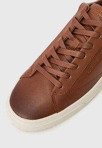 Zapatillas de cuero marrón con cordones planos marrones, puntera redondeada, suela de goma blanca y logotipo grabado en el lateral.