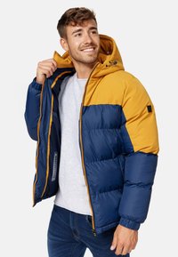 Polstret jakke i marineblå og sennepsgul. Har hætte, lynlås med orange detaljer og elastiske manchetter. Tekstureret overflade med et slankt design.