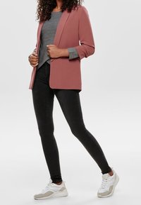 Blazer rosa blush con spalle strutturate, top grigio, jeans skinny neri e sneaker bianche con dettagli grigi. Texture lisce in tutto.