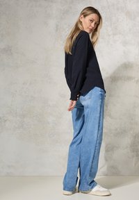 Marineblauwe blouse met lange mouwen, gemaakt van textuurstof, met knoopaccenten, gecombineerd met lichtblauwe wijde jeans en witte sneakers op een neutrale achtergrond.