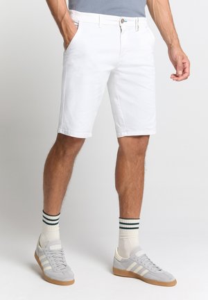 Weiße Baumwollshorts mit Fronttaschen, etwas oberhalb des Knies, kombiniert mit grauen Turnschuhen und gestreiften Socken.