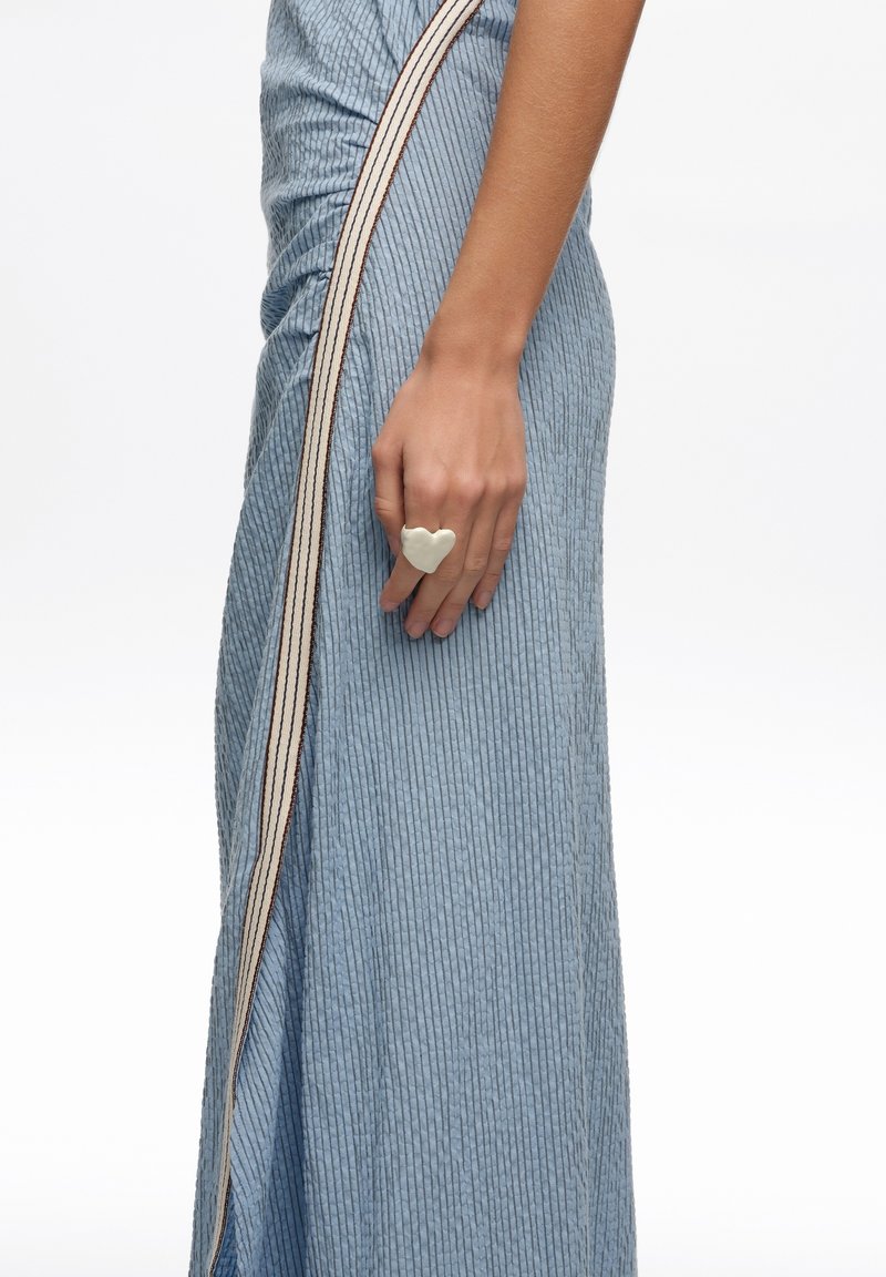 Mujer con una falda azul texturizada con rayas verticales y un gran anillo blanco en forma de corazón en el dedo, con un fondo liso.