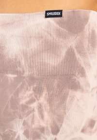 Lichtroze geribbelde stof met een tie-dye patroon, voorzien van een zwart label met "SMILODOX" aan de bovenrand. Zachte textuur, nauwsluitende pasvorm.