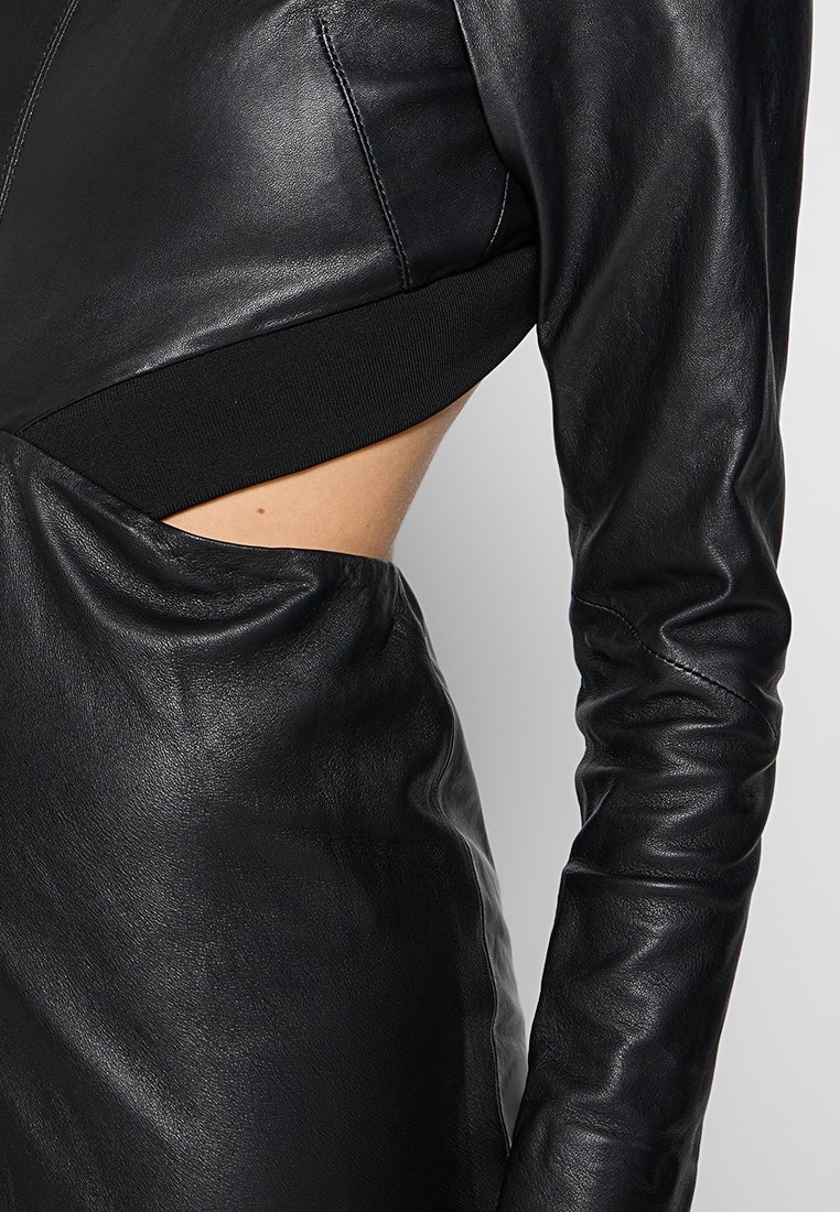 Zbliżenie na czarną skórzaną sukienkę z długim rękawem, z bocznym wycięciem odsłaniającym skórę oraz prążkowanym czarnym pasem.