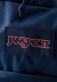 Κοντινή λήψη ύφασματος σάκιδιου πλάτης JanSport σε ναυτικό μπλε με ραμμένο πατς που φέρει το λογότυπο της JanSport σε κόκκινο και άσπρο.