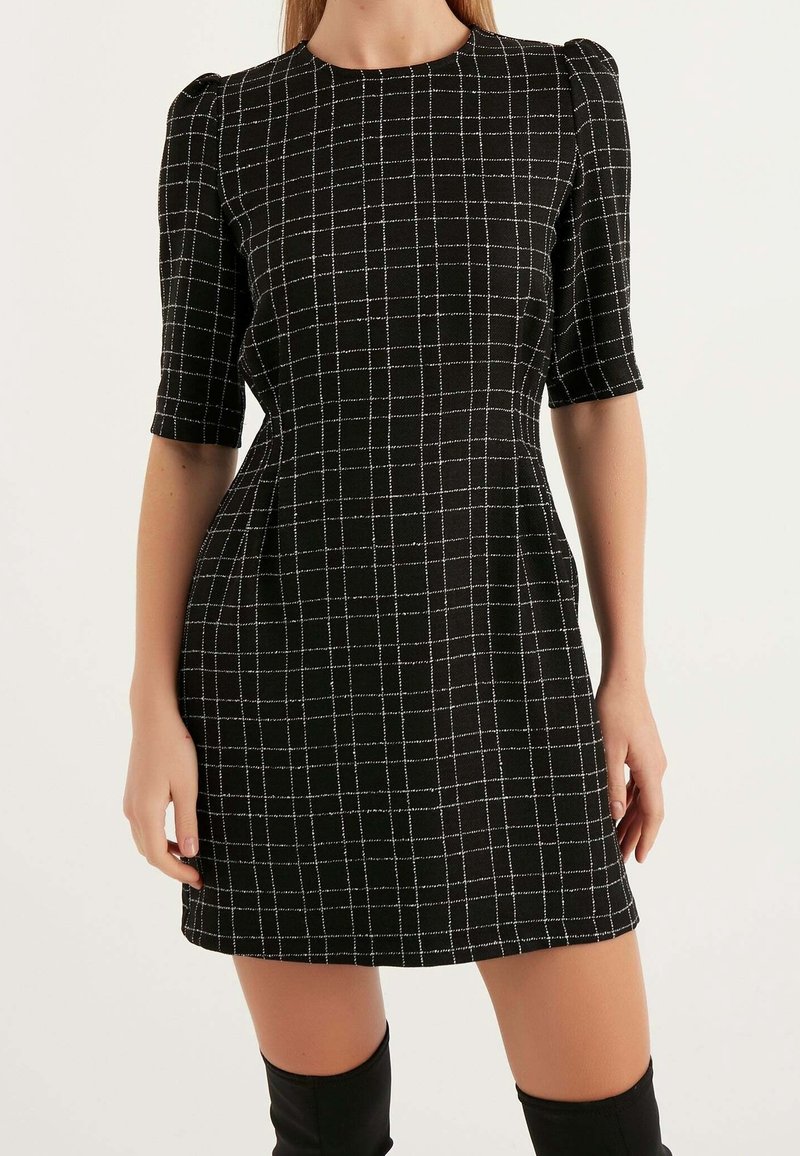 Robe mini noire avec motif à carreaux blancs, manches courtes bouffantes et taille cintrée, portée avec des bottes noires montantes au genou.