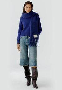 Pull marin, écharpe assortie, jean large bleu clair, et bottes marron jusqu'au genou. L'écharpe présente des franges et une étiquette de marque.