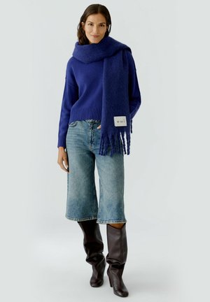Pull marin, écharpe assortie, jean large bleu clair, et bottes marron jusqu'au genou. L'écharpe présente des franges et une étiquette de marque.