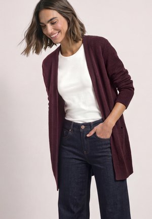 Femme souriante portant une chemise blanche, un jean bleu foncé et un long cardigan bordeaux avec des poches, debout avec une main dans sa poche.